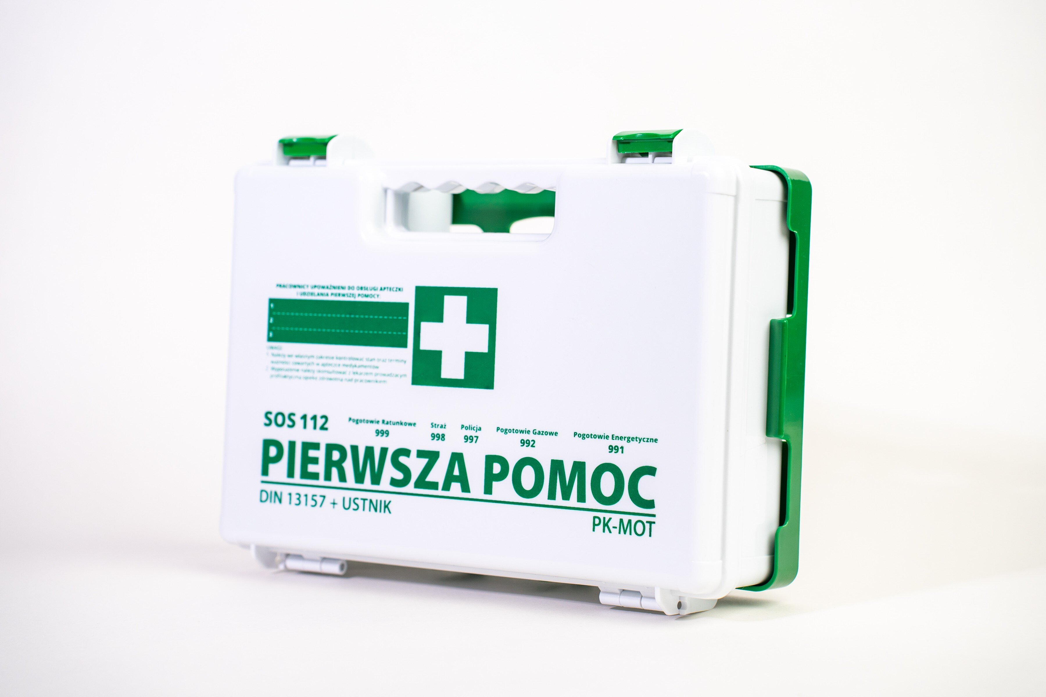 Biała apteczka z zielonymi elementami i uchwytem, na której widnieje napis \'PIERWSZA POMOC\' oraz symbol krzyża. Dodatkowo widoczne są numery alarmowe i oznaczenie \'DIN 13157 + USTNIK\'.