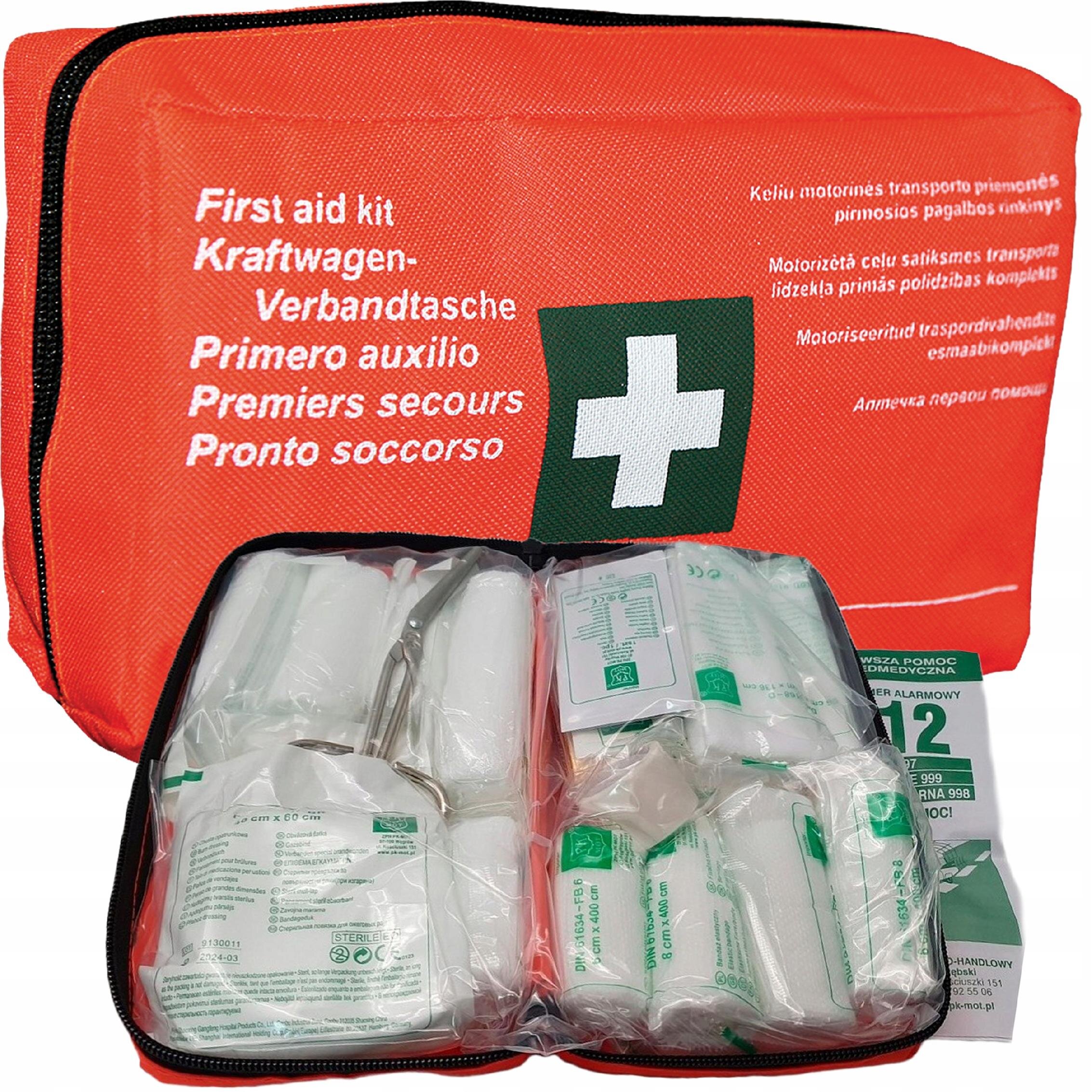 Czerwona apteczka pierwszej pomocy z białym krzyżem na zielonym tle, zawierająca różne materiały opatrunkowe. Na apteczce widoczny jest tekst: \'First aid kit, Kraftwagen-Verbandtasche, Primero auxilio, Premiers secours, Pronto soccorso\'.