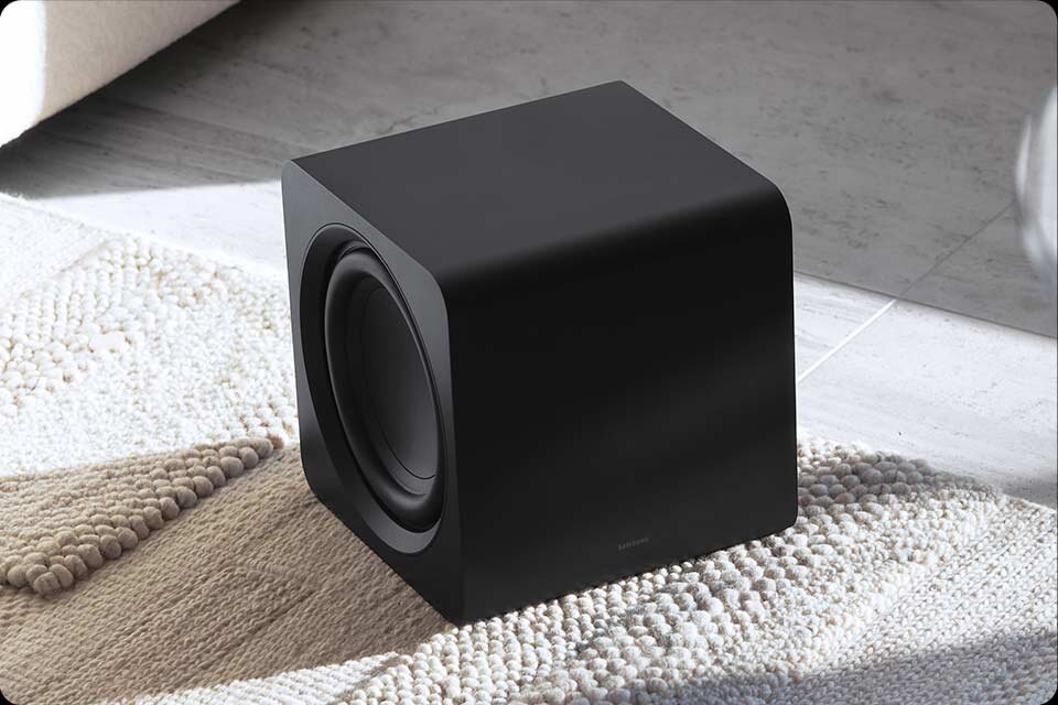 Czarny subwoofer o prostokątnym kształcie stoi na jasnym, teksturowanym dywanie. Na przedniej części urządzenia widoczne jest logo \'SAMSUNG\'.