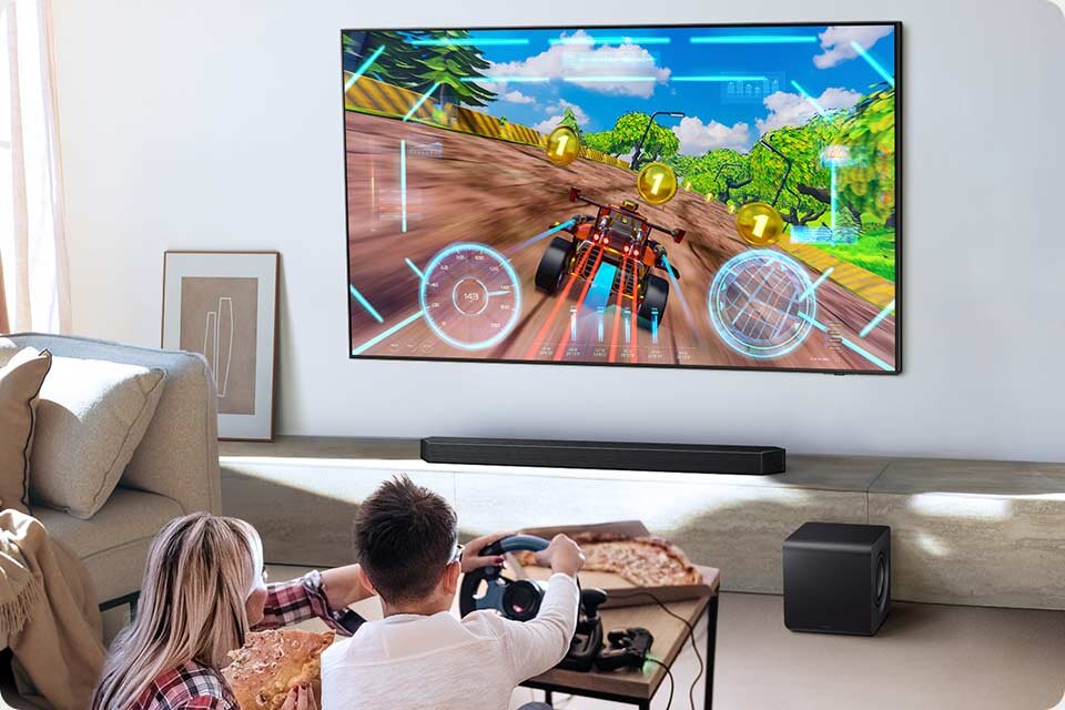 Na ścianie wisi duży telewizor, na którym wyświetlana jest gra wyścigowa. Przed telewizorem znajduje się soundbar i subwoofer, a dwie osoby siedzą na kanapie, grając w grę.