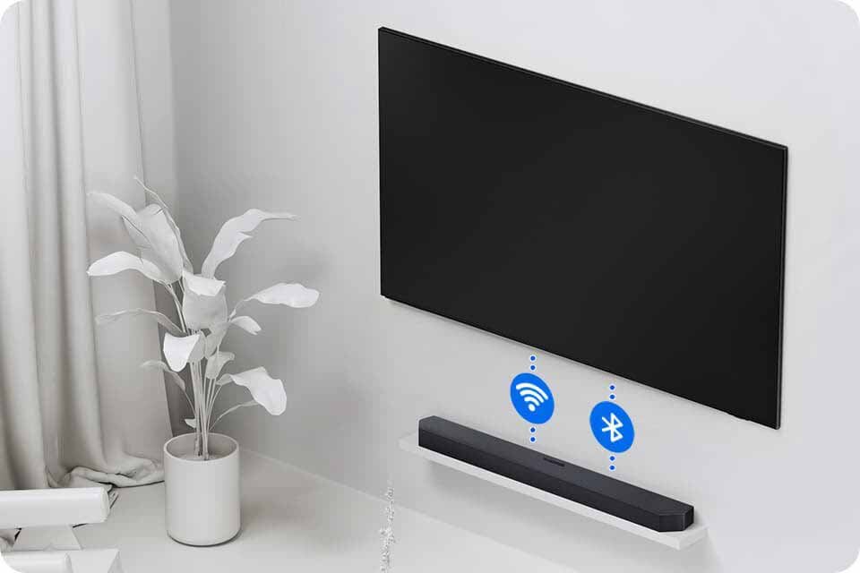 Na ścianie wisi duży, czarny telewizor, a poniżej na półce znajduje się czarny soundbar. Obok soundbara widoczne są ikony Wi-Fi i Bluetooth.