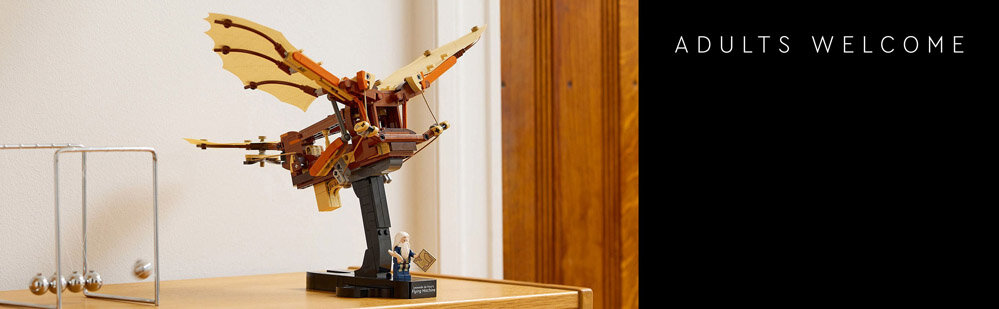 Na drewnianym stole stoi model maszyny latającej z klocków LEGO, obok znajduje się wahadło Newtona. Po prawej stronie na czarnym tle widnieje napis \'ADULTS WELCOME\'.