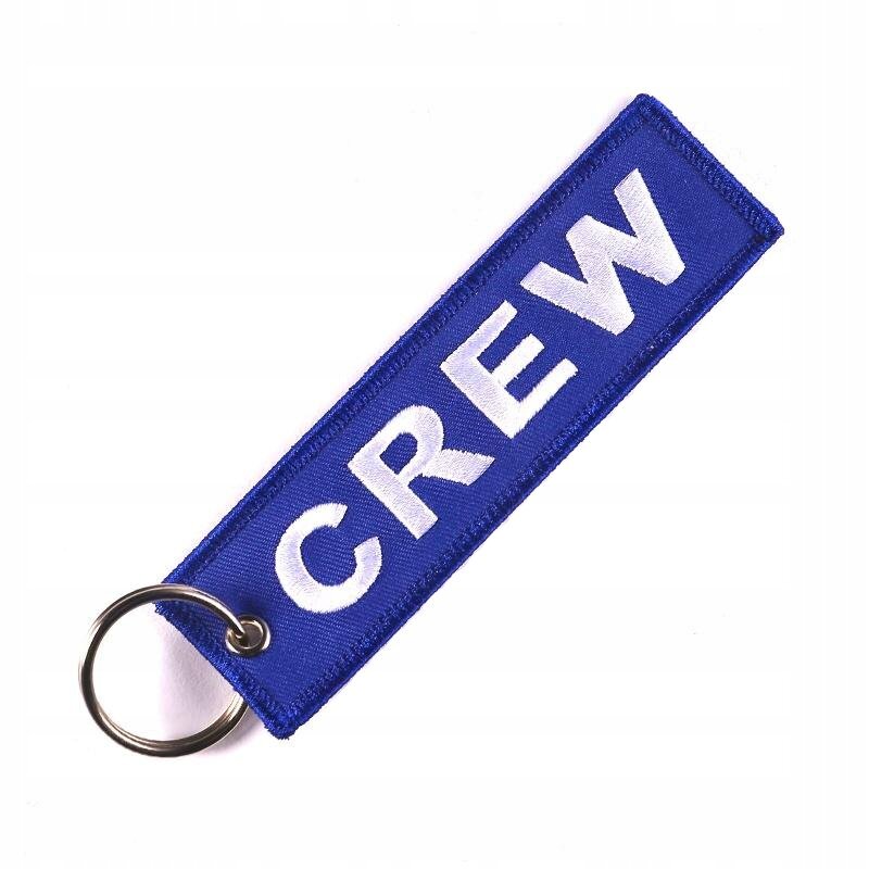 Niebieski brelok z napisem \'CREW\' w białym kolorze, posiada metalowe kółko do zawieszania.