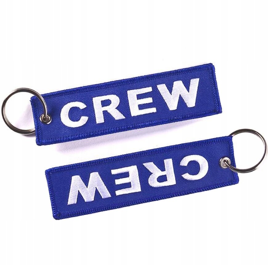 Na obrazie widoczne są dwa niebieskie breloki z napisem \'CREW\' w białym kolorze. Każdy brelok ma metalowe kółko do zawieszenia kluczy.