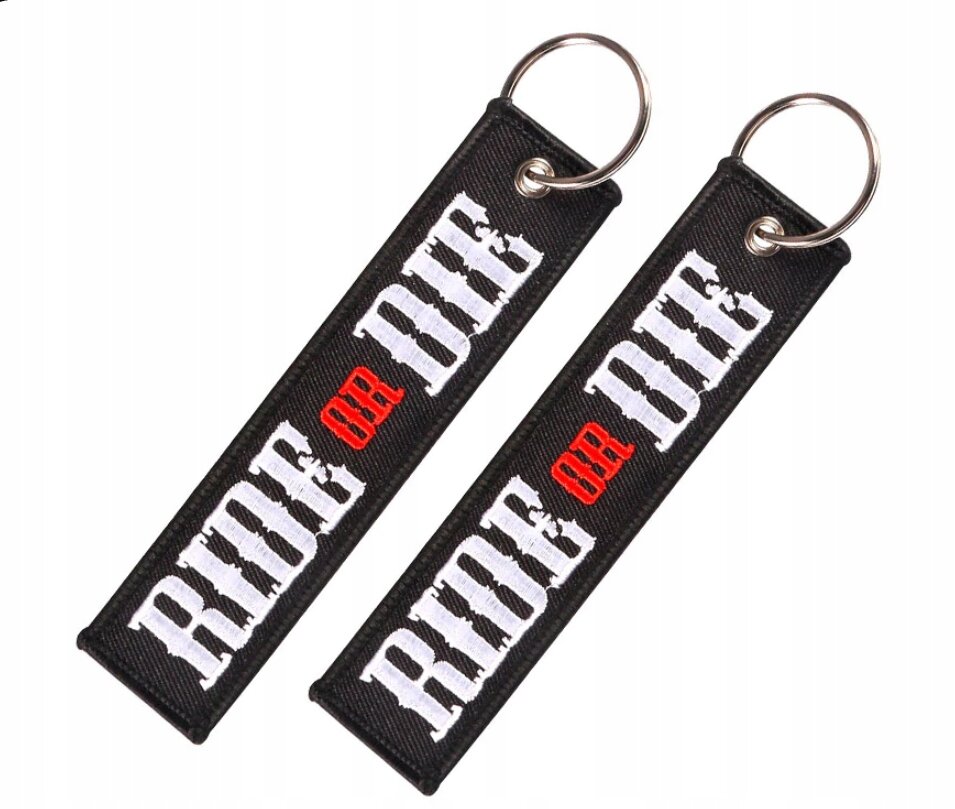 Dwa czarne breloki z napisem \'RIDE OR DIE\', gdzie \'OR\' jest w kolorze czerwonym. Każdy brelok ma metalowe kółko do zawieszenia kluczy.