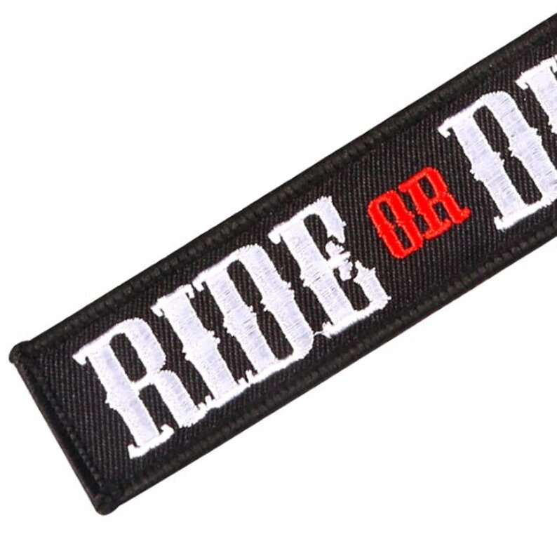 Czarny brelok z białym napisem \'RIDE\' i \'DIE\', oraz czerwonym \'OR\'.