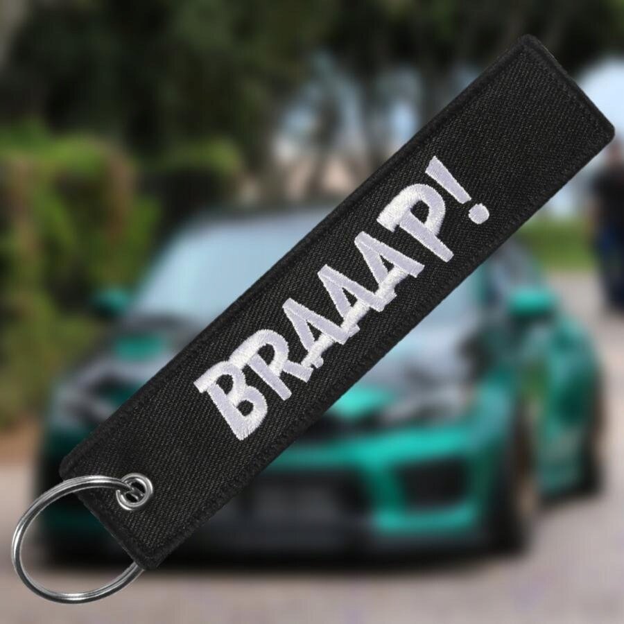 Czarny brelok z napisem \'BRAAAP!\' w białym kolorze, z metalowym kółkiem do zawieszania. W tle rozmyty obraz samochodu.