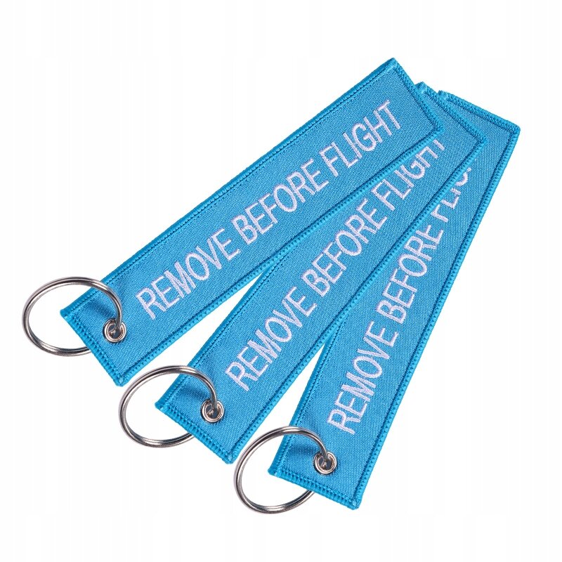 Na obrazie widoczne są trzy niebieskie breloki z napisem \'REMOVE BEFORE FLIGHT\', każdy z metalowym kółkiem do zawieszenia.