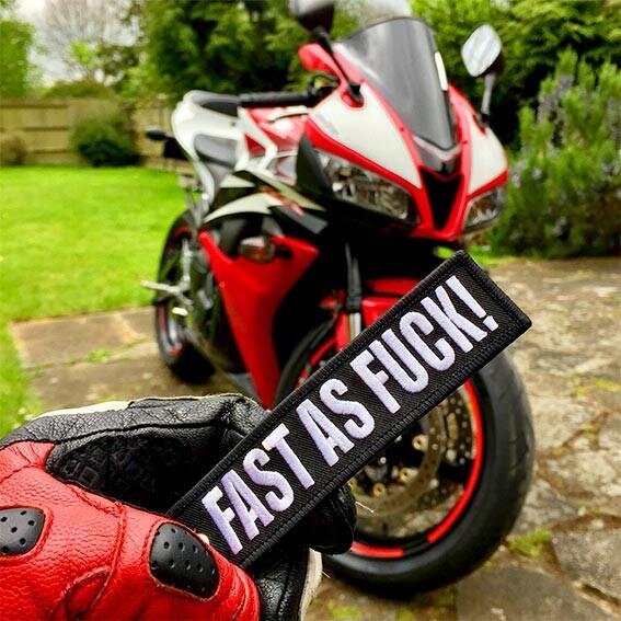 Osoba w rękawicy motocyklowej trzyma brelok z napisem \'FAST AS FUCK!\'. W tle widoczny jest motocykl w kolorach czerwonym, czarnym i białym, stojący na podjeździe obok trawnika.