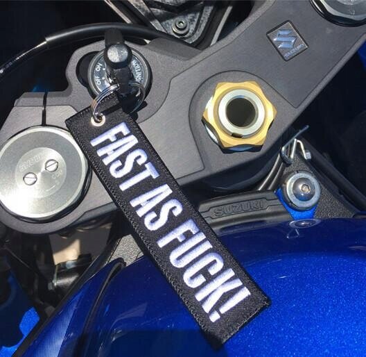 Na zdjęciu widoczny jest brelok z napisem \'FAST AS FUCK!\' przymocowany do kluczyków motocykla. W tle widać elementy motocykla oraz logo Suzuki.