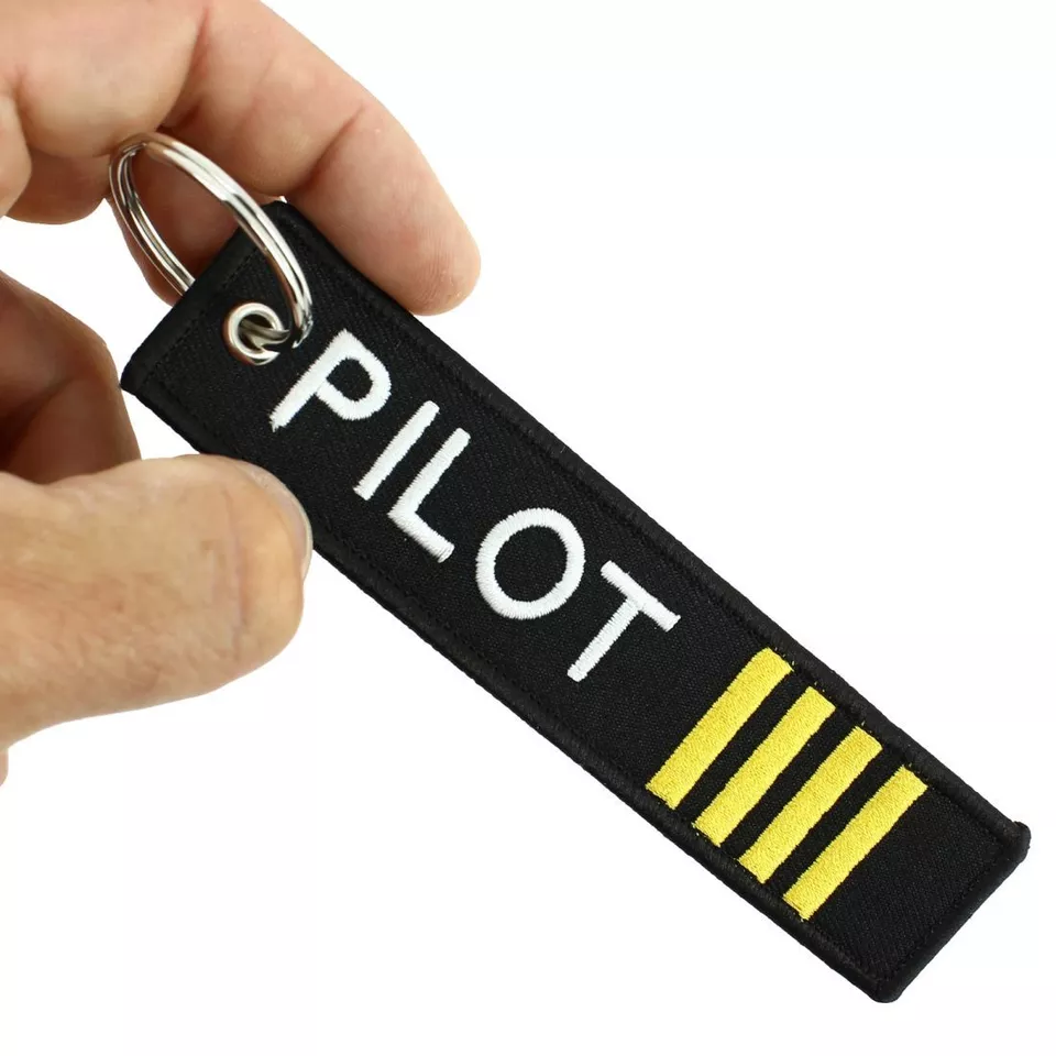 Czarny brelok do kluczy z napisem \'PILOT\' w białym kolorze oraz trzema żółtymi paskami. Brelok trzymany jest w dłoni i posiada metalowe kółko do zawieszenia.