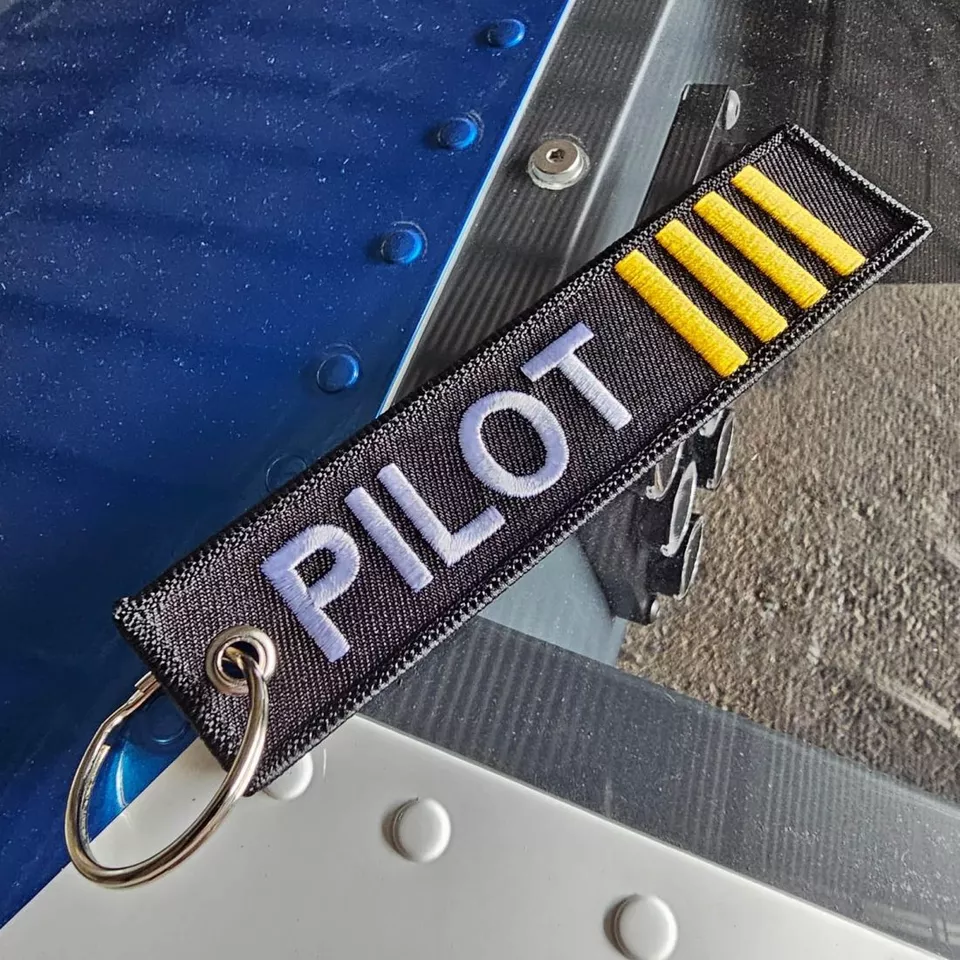Na zdjęciu widoczny jest brelok do kluczy z napisem \'PILOT\' oraz trzema żółtymi paskami. Brelok leży na metalowej powierzchni z nitami.