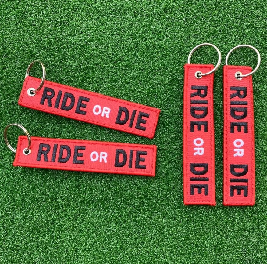 Na zielonym tle znajdują się cztery czerwone breloki z napisem \'RIDE or DIE\'. Każdy brelok ma metalowe kółko do zawieszania.