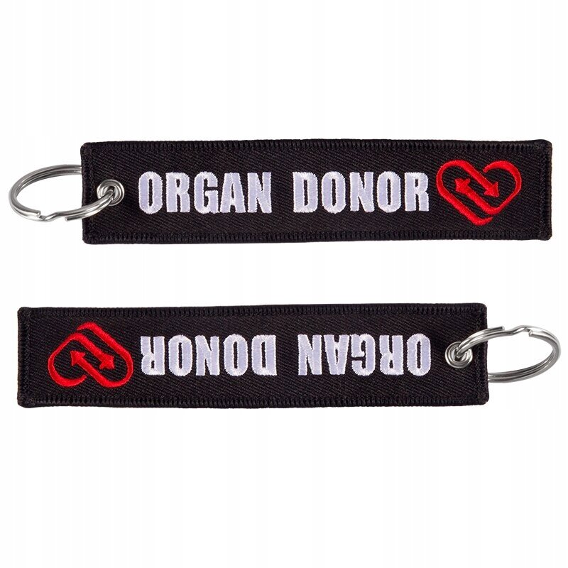 Na obrazie widoczne są dwa czarne breloki z napisem \'ORGAN DONOR\' w białym kolorze oraz czerwonym symbolem serca z dwiema strzałkami. Każdy brelok ma metalowe kółko do zawieszenia kluczy.