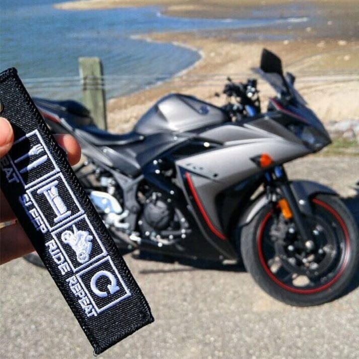Na pierwszym planie widoczny jest czarny brelok z białymi symbolami i napisem \'EAT SLEEP RIDE REPEAT\'. W tle znajduje się szary motocykl zaparkowany na tle plaży i wody.