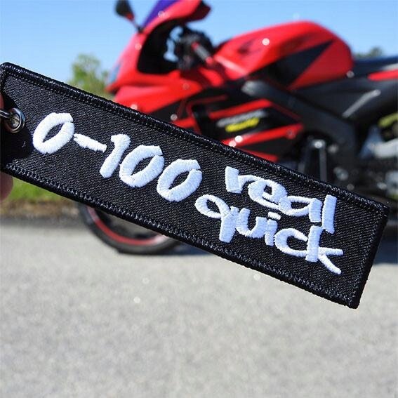 Na pierwszym planie widoczny jest czarny brelok z białym napisem \'0-100 real quick\'. W tle znajduje się czerwony motocykl.