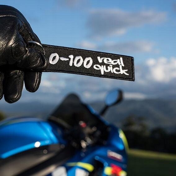 Czarna zawieszka do kluczy z białym napisem \'0-100 real quick\' trzymana w rękawicy motocyklowej. W tle widoczny jest niebieski motocykl i krajobraz z górami.