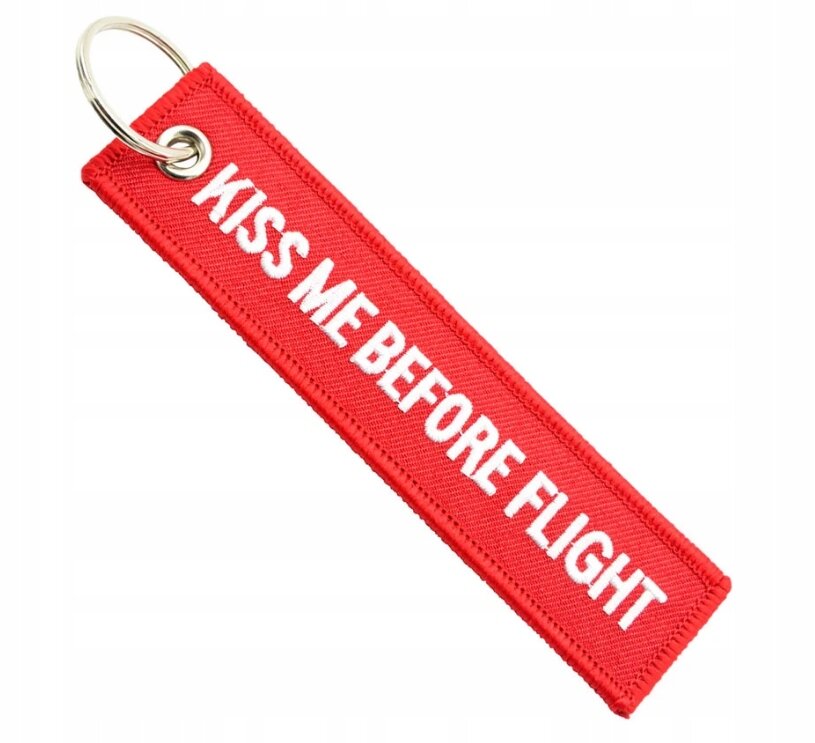 Czerwony brelok z napisem \'KISS ME BEFORE FLIGHT\' w białym kolorze, z metalowym kółkiem do zawieszenia.
