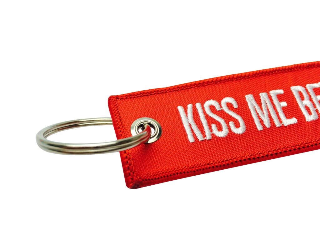 Czerwony brelok z metalowym kółkiem i białym napisem \'KISS ME BEFORE FLIGHT\'.