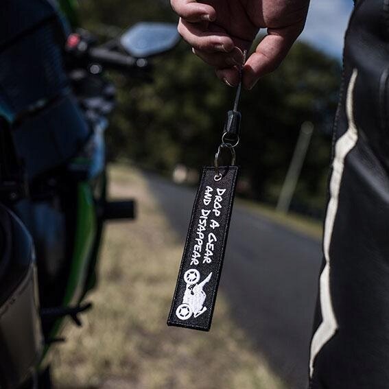 Osoba trzyma brelok z napisem \'DROP A GEAR AND DISAPPEAR\' oraz grafiką motocykla. W tle widać rozmytą drogę i fragment motocykla.