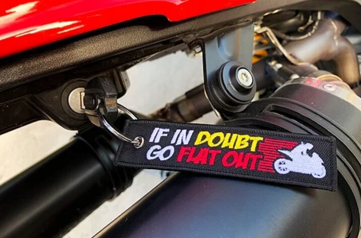 Czarny brelok z napisem \'IF IN DOUBT GO FLAT OUT\' w kolorach białym, żółtym i czerwonym oraz grafiką motocykla, przymocowany do klucza w stacyjce pojazdu.