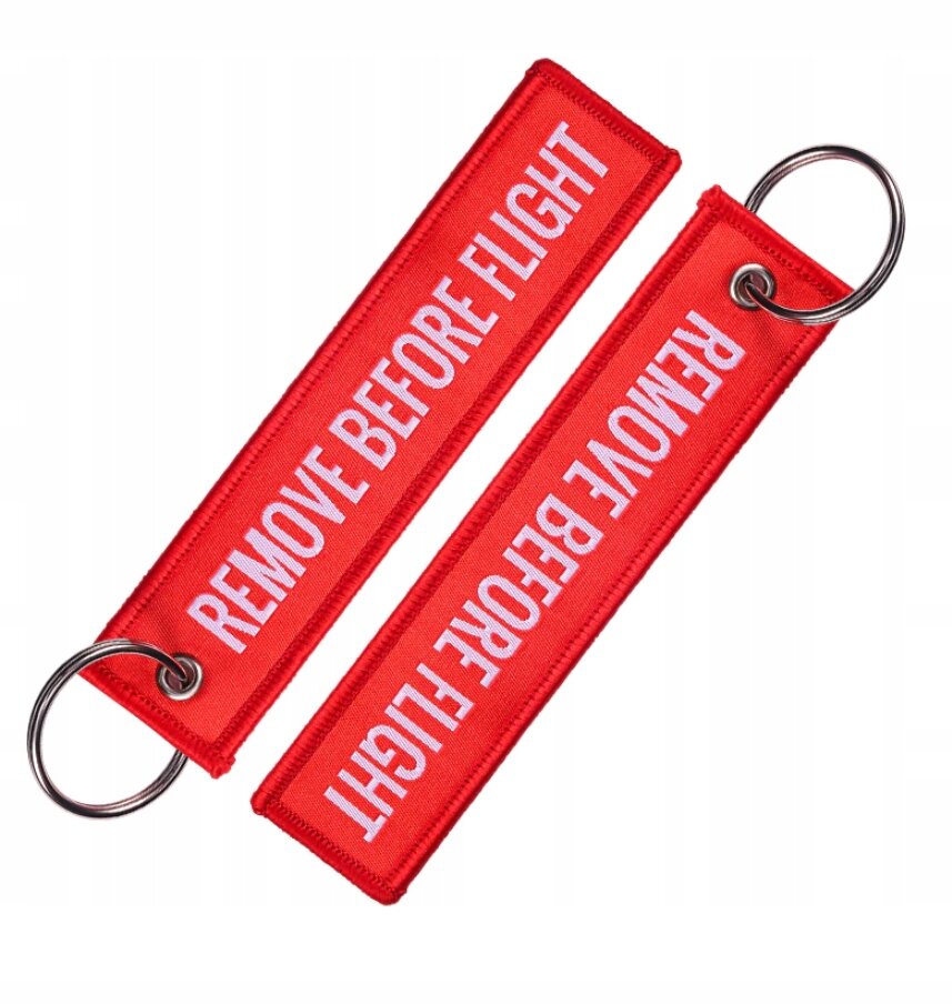 Dwa czerwone breloki z białym napisem \'REMOVE BEFORE FLIGHT\', każdy z metalowym kółkiem do zawieszenia.