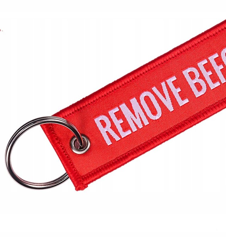 Czerwony brelok z napisem \'REMOVE BEFORE FLIGHT\' wykonany z materiału, z metalowym kółkiem do kluczy.