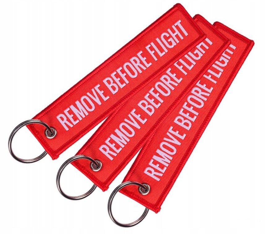 Trzy czerwone breloki z napisem \'REMOVE BEFORE FLIGHT\' w białym kolorze, każdy z metalowym kółkiem do kluczy.