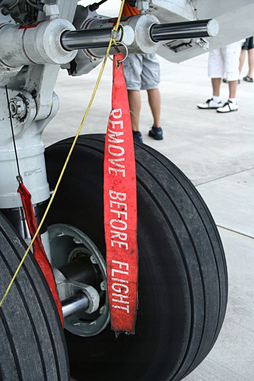 Czerwony brelok z napisem \'REMOVE BEFORE FLIGHT\' jest przymocowany do podwozia samolotu. W tle widoczne są koła i fragmenty samolotu oraz nogi kilku osób.