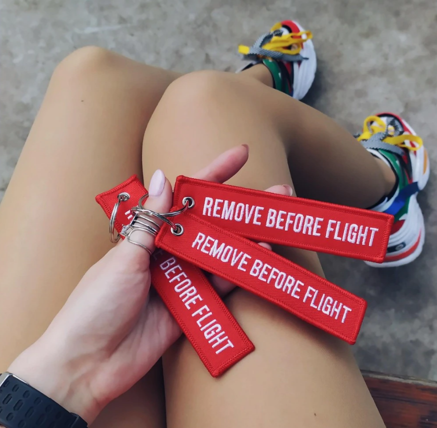 Osoba trzyma trzy czerwone breloki z napisem \'REMOVE BEFORE FLIGHT\'. W tle widoczne są nogi i kolorowe buty sportowe.