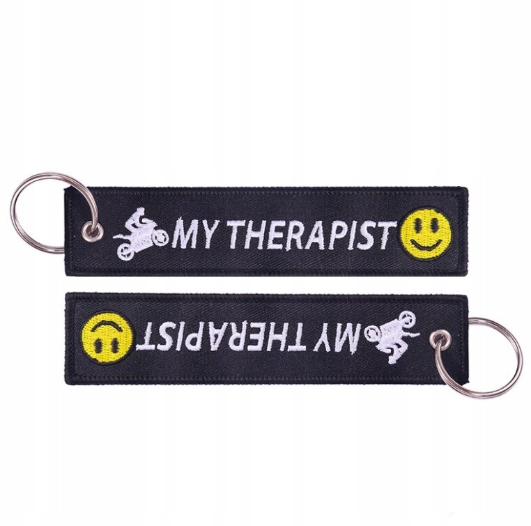 Czarny brelok z napisem \'MY THERAPIST\' oraz żółtą emotikoną uśmiechu i białym symbolem motocyklisty. Brelok posiada metalowe kółko do zawieszenia kluczy.