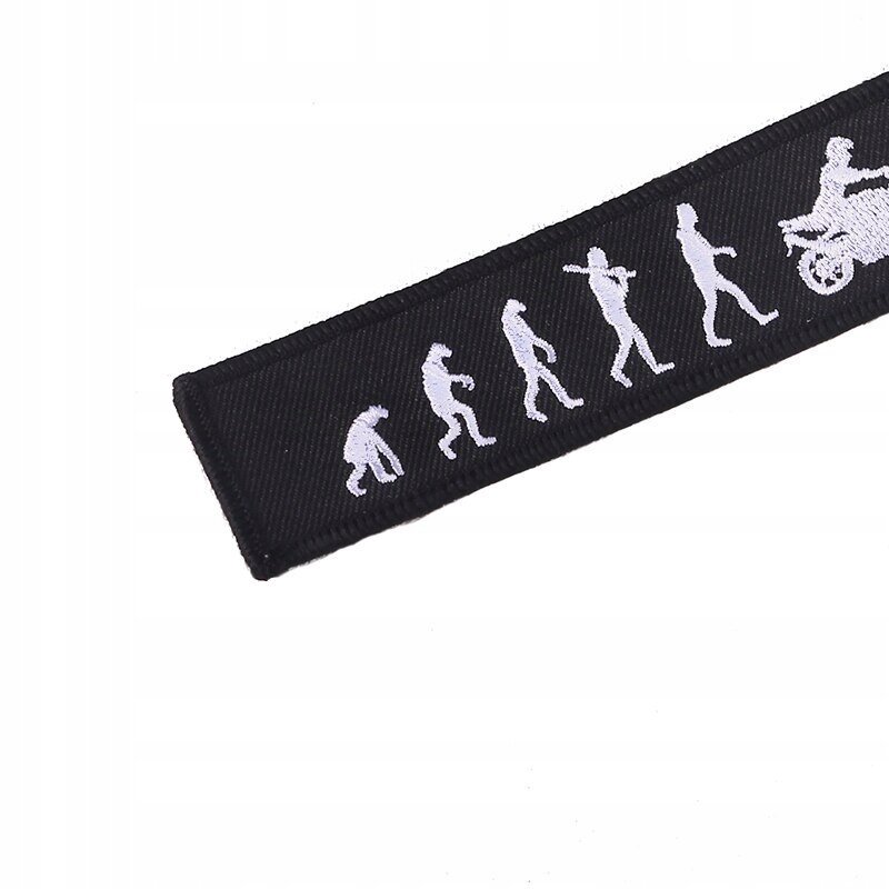 Czarny brelok do kluczy z białymi sylwetkami przedstawiającymi ewolucję człowieka, kończącą się postacią na motocyklu.