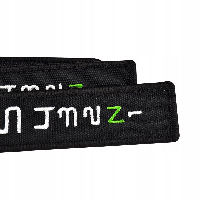 Na czarnym tle widoczne są białe i zielone haftowane symbole przypominające litery i cyfry. Produkt ma formę prostokątnej naszywki z obszytymi krawędziami.