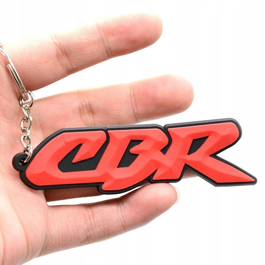 Czerwony brelok do kluczy z napisem \'CBR\' trzymany w dłoni. Brelok ma czarne obramowanie i jest przymocowany do metalowego łańcuszka.