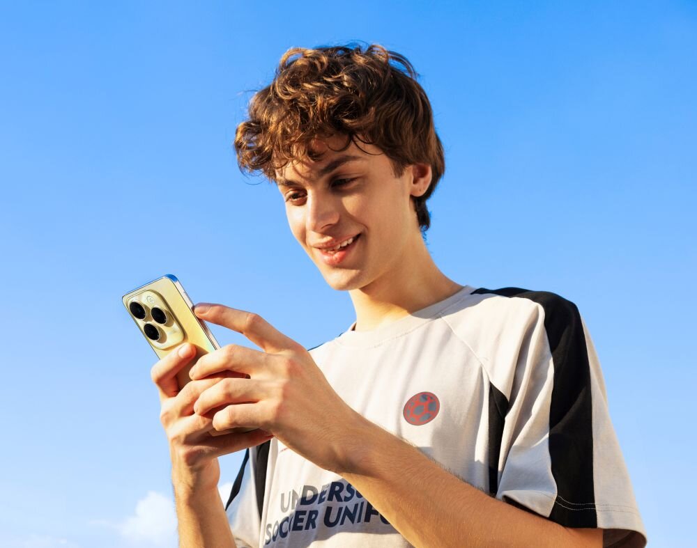 Osoba trzyma smartfon z trzema dużymi obiektywami aparatu. W tle widać jasne, niebieskie niebo.