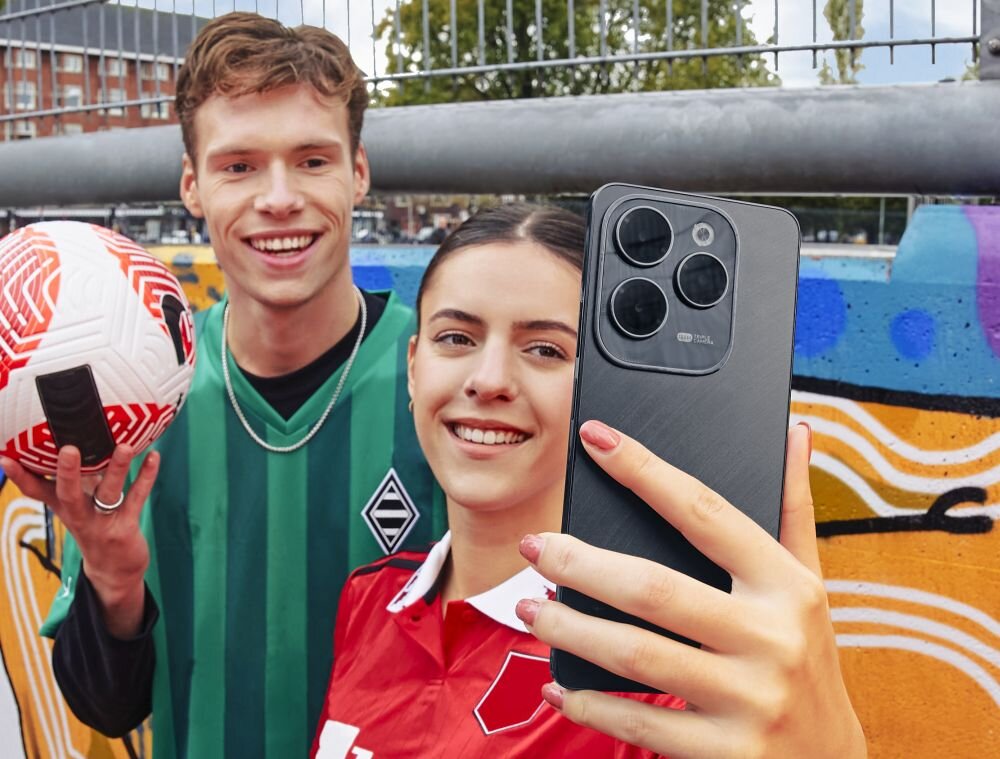 Dwie osoby robią selfie smartfonem z trzema dużymi obiektywami aparatu. W tle widać kolorowy mural i fragment boiska sportowego.