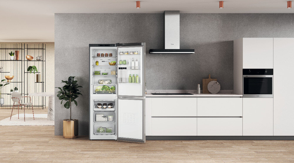 Kuchnia z otwartą lodówką w kolorze srebrnym, wypełnioną różnymi produktami spożywczymi. Obok znajduje się nowoczesna zabudowa kuchenna z piekarnikiem i okapem.
