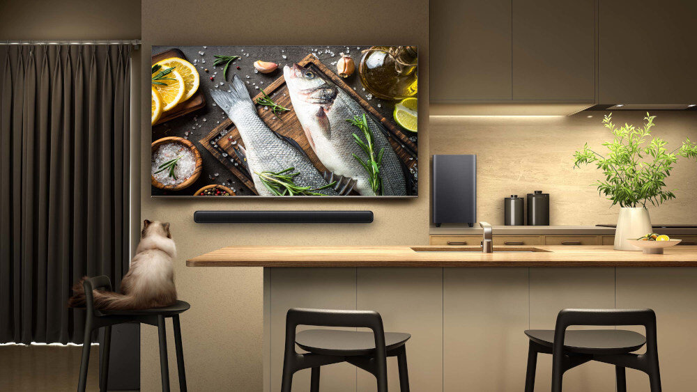 W nowoczesnej kuchni na ścianie wisi duży telewizor z obrazem ryb i cytryn, pod którym znajduje się czarny soundbar. Na blacie siedzi kot, a obok stoi wazon z zieloną rośliną.