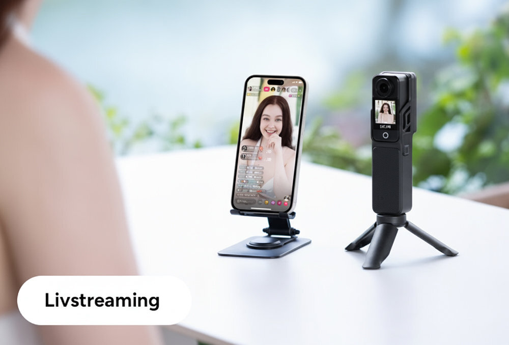 Na stole znajduje się smartfon na stojaku, wyświetlający transmisję na żywo z osobą oraz kamera sportowa na małym statywie. Na obrazie widoczny jest napis \'Livstreaming\'.