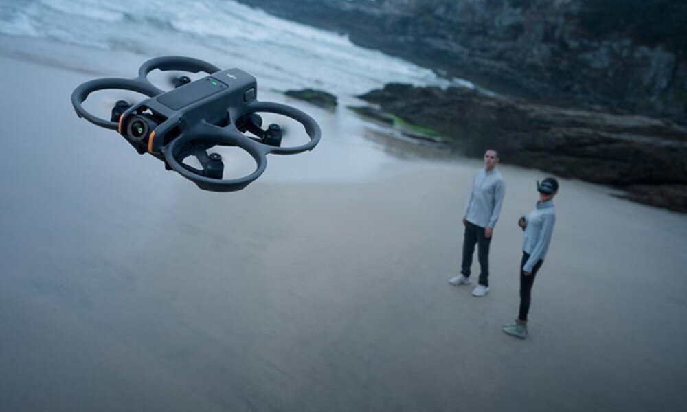 Dron unosi się nad piaszczystą plażą, w tle widać dwie osoby stojące na piasku. W tle znajdują się skały i fale oceanu.