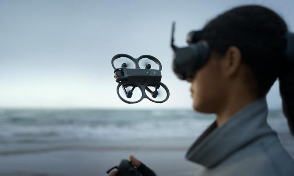 Osoba w goglach VR kontroluje drona unoszącego się w powietrzu na tle morza. Dron ma kompaktową konstrukcję z osłonami na śmigła.