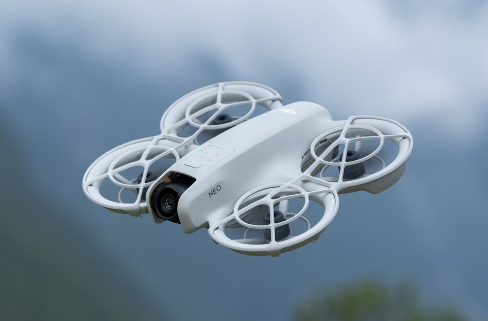 Na obrazie widoczny jest biały dron z czterema osłoniętymi śmigłami, unoszący się w powietrzu na tle górskiego krajobrazu. Na dronie znajduje się napis \'NEO\'.