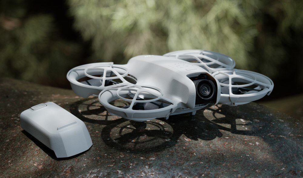 Na obrazie widoczny jest biały dron z osłonami na śmigła, stojący na kamiennej powierzchni. Obok drona leży dodatkowa bateria.