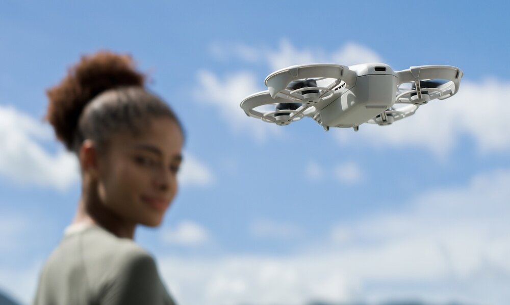 Na tle nieba widoczny jest biały dron z osłonami na śmigła. W tle rozmyta postać osoby.