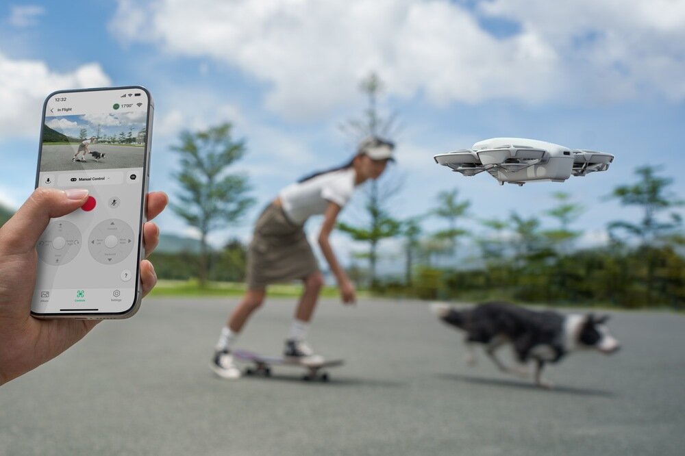 Osoba trzyma smartfon z aplikacją do sterowania dronem, który unosi się w powietrzu. W tle widać osobę na deskorolce i psa biegnącego po asfalcie.