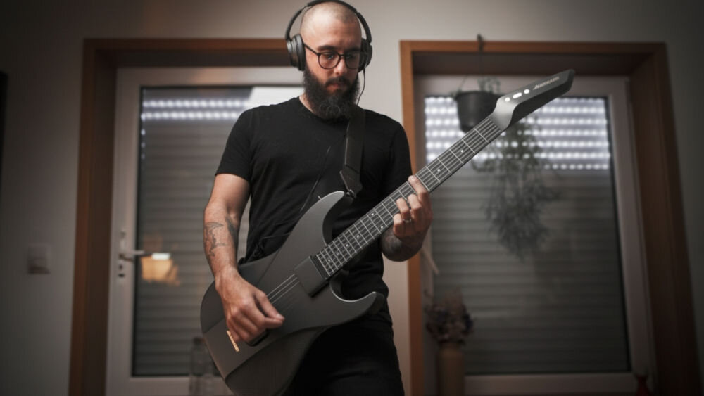 Osoba gra na czarnej gitarze elektrycznej, nosząc słuchawki. W tle widoczne są drzwi z żaluzjami i roślina.
