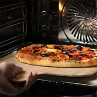 Wnętrze piekarnika z widoczną pizzą na drewnianej łopatce. W tle widać wentylator piekarnika.