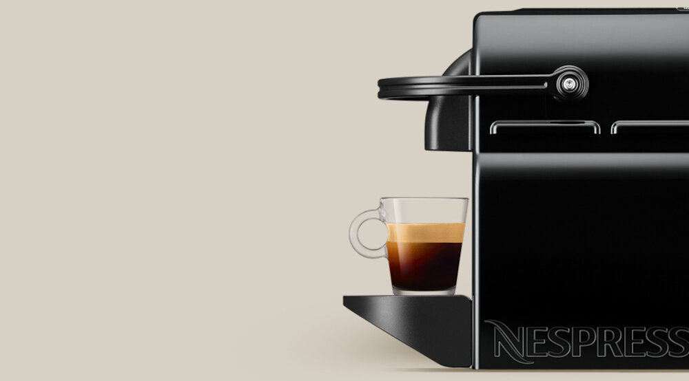 Czarny ekspres do kawy z filiżanką espresso na podstawce. Na obudowie widoczne jest logo \'NESPRESSO\'.