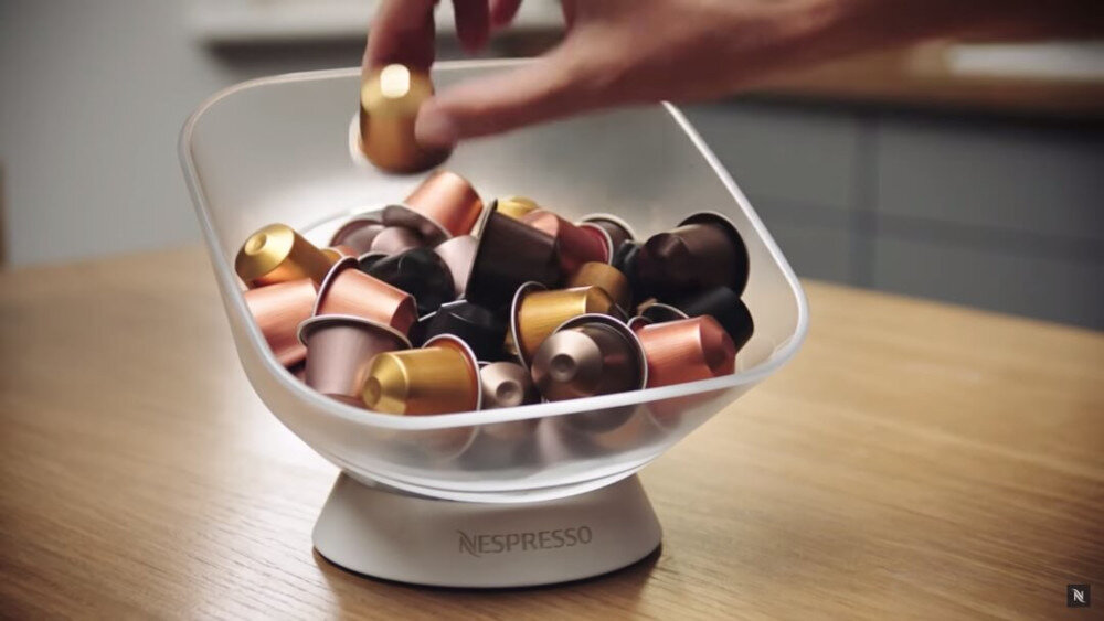 Przezroczysta miska wypełniona kolorowymi kapsułkami do kawy, z logo \'Nespresso\' na podstawie. Ręka sięga po jedną z kapsułek.