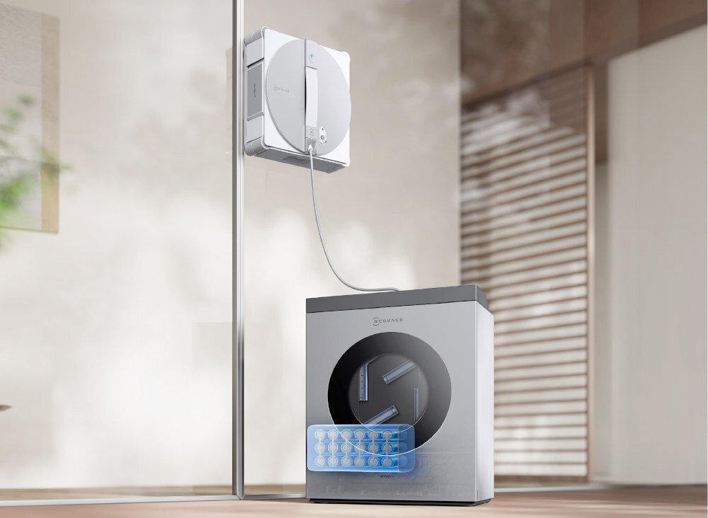 Na szklanej powierzchni znajduje się robot do mycia okien podłączony przewodem do urządzenia stojącego na podłodze. Na urządzeniu widoczne jest logo \'ECOVACS\'.